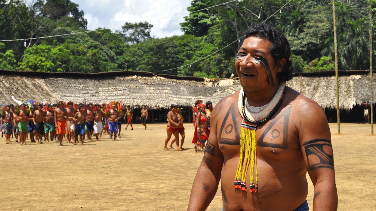 En Yanomami-mann med tradisjonelle klær og ansiktsmaling smiler inn i kamera, mens mange andre står bak ham.